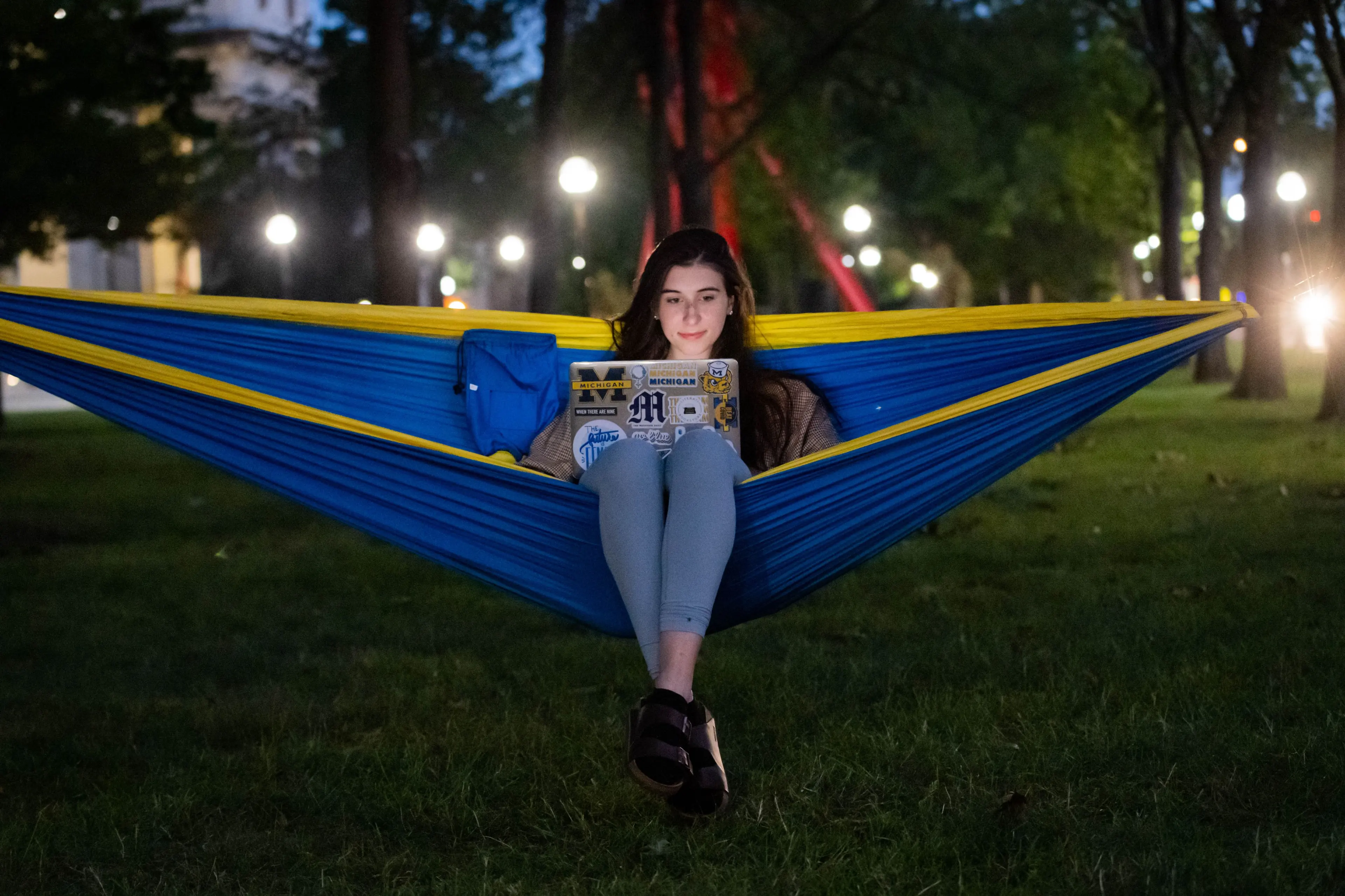 A female student with long brown hair, relaxes in a maize and blue hammock while reading on her laptop.